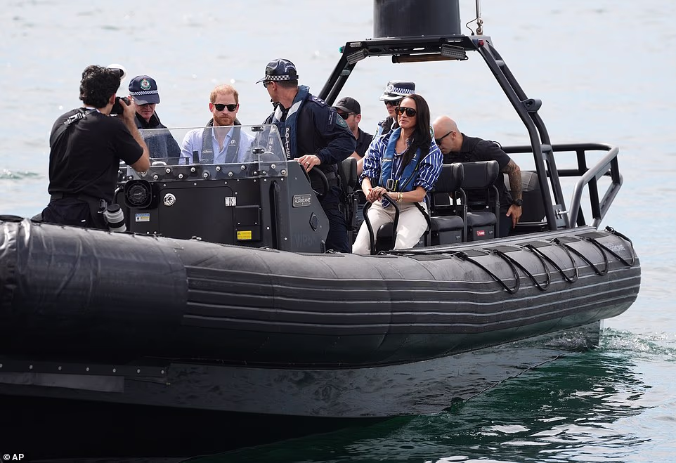 Before the money-spinning commercial event, the Sussexes took in the delights of Sydney on the final day of their Australian tour. They sailed around the world-famous Sydney Harbour after a police escort and posed for selfies with fans at the Sydney Opera House at an Invictus Australia event. After he looked angry yesterday when asked for a selfie by someone blocking Meghan's path, today Harry moved away a camera crew who came too close. Harry and Meghan looked much happier when they were gifted matching custom-made pairs of Invictus Australia-branded flip-flops, known as thongs Down Under. Harry's were emblazoned with 'G'day Hazza' while Meghan's had 'G'day Megs'. Some of the group they spoke to were among the first responders to the Bondi terrorist attack, in which 15 innocent lives were lost on December 14. The Duke and Duchess of Sussex looked deeply moved and chatted to Jessica Chapnik Kahn, who survived while shielding her five-year-old daughter after attending a Hanukkah party. Ms Chapnik Kahn, who was embraced by Meghan, said it was an 'honour' to meet the couple during their visit to the Bondi Surf Bathers' Life Saving Club. She said: 'The greater problem in the world right now is hate.