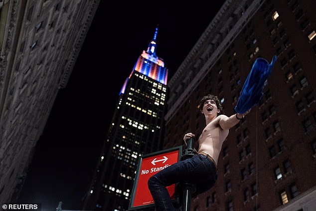 A shirtless New Yorker swings from a sign post with the Empire State Building in the back
